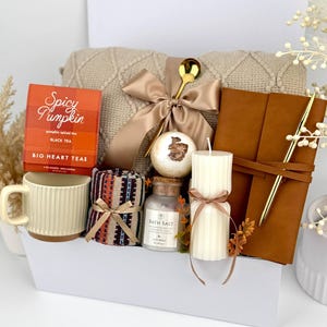 May include: A gift box containing a beige knit blanket with a satin bow, a box of Spicy Pumpkin black tea, a ribbed cream mug, a patterned fabric roll, a bath bomb, a glass jar of bath salts, a ribbed pillar candle, and a brown leather journal with a gold pen.