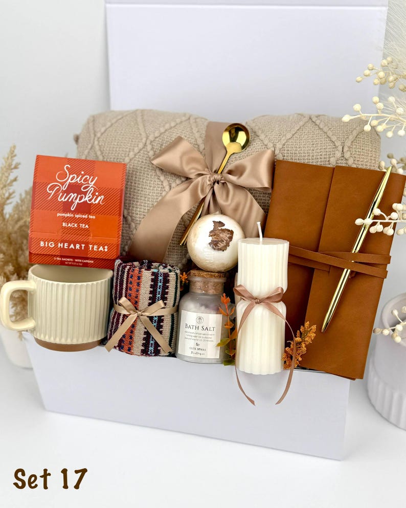 May include: A gift box containing a beige knit blanket with a satin bow, a box of Spicy Pumpkin black tea, a ribbed cream mug, a patterned fabric roll, a bath bomb, a glass jar of bath salts, a ribbed pillar candle, and a brown leather journal with a gold pen.