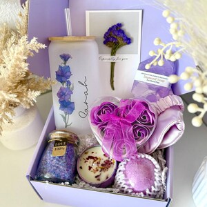 May include: A lilac gift box containing self-care products. It features a glass tumbler with a floral pattern, a candle, bath salts, a heart-shaped box of rose soaps, a sleep mask, and a soap bar. The box has the text "Set 8".