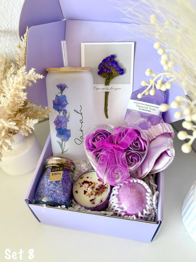 May include: A lilac gift box containing self-care products. It features a glass tumbler with a floral pattern, a candle, bath salts, a heart-shaped box of rose soaps, a sleep mask, and a soap bar. The box has the text "Set 8".