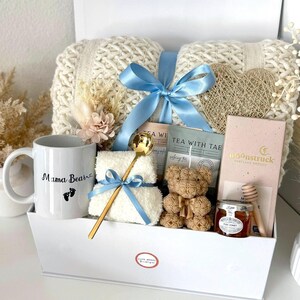K&ouml;nnte beinhalten: Eine wei&szlig;e Geschenkbox gef&uuml;llt mit einer cremefarbenen Strickdecke, einer Tasse mit dem Text "Mama Bear", einem Teddyb&auml;ren, Tee, Honig und einem goldenen L&ouml;ffel. Die Box ist mit einem hellblauen Band gebunden. Trockenblumen und eine herzf&ouml;rmige Dekoration sind ebenfalls enthalten.