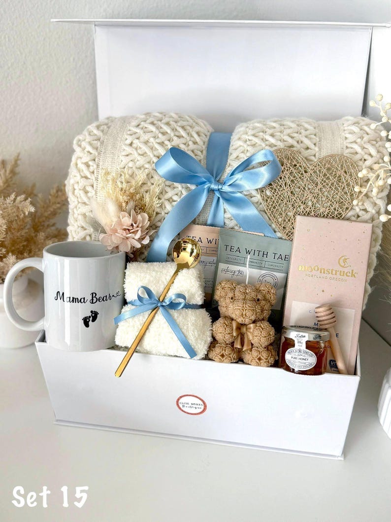 K&ouml;nnte beinhalten: Eine wei&szlig;e Geschenkbox gef&uuml;llt mit einer cremefarbenen Strickdecke, einer Tasse mit dem Text "Mama Bear", einem Teddyb&auml;ren, Tee, Honig und einem goldenen L&ouml;ffel. Die Box ist mit einem hellblauen Band gebunden. Trockenblumen und eine herzf&ouml;rmige Dekoration sind ebenfalls enthalten.