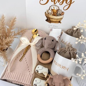 May include: A gift box labeled Hello Baby containing a pink knit blanket with a cream ribbon, a gold spoon, a crocheted elephant rattle with a wooden ring, two white mugs labeled daddy and mommy, and a brown elephant-shaped candle.