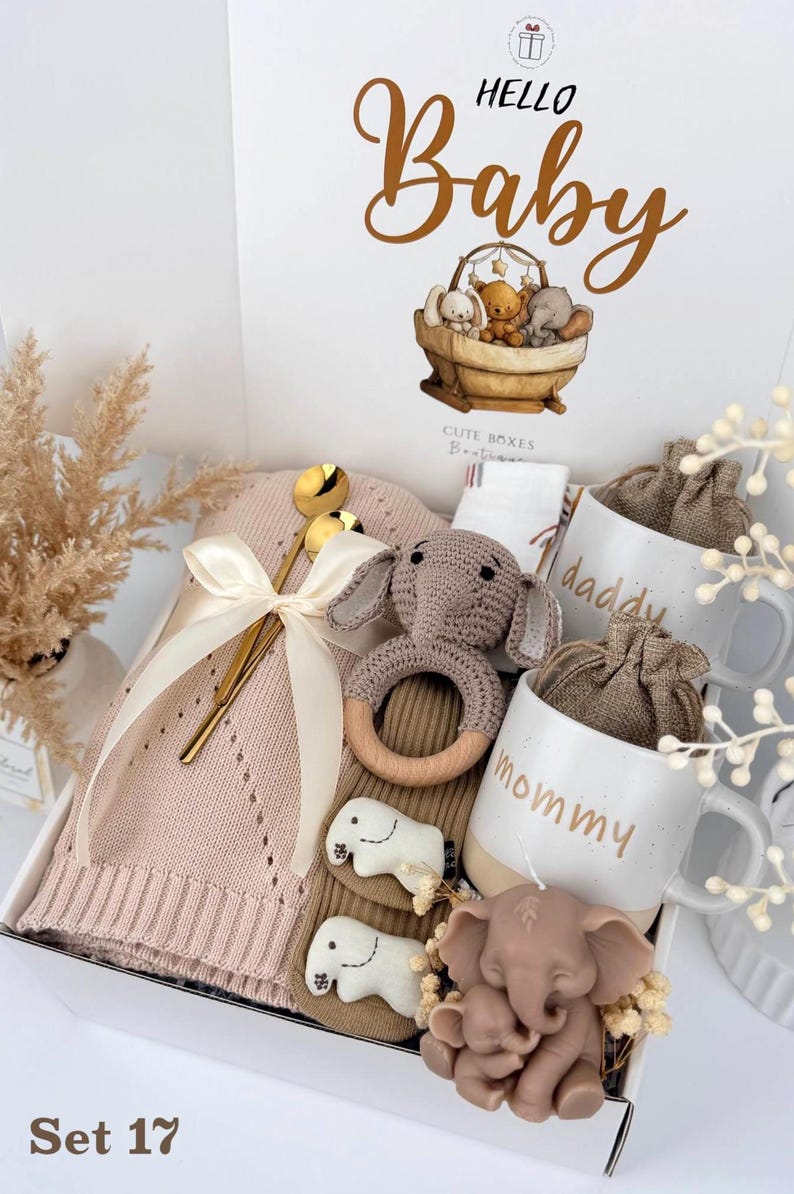 May include: A gift box labeled Hello Baby containing a pink knit blanket with a cream ribbon, a gold spoon, a crocheted elephant rattle with a wooden ring, two white mugs labeled daddy and mommy, and a brown elephant-shaped candle.