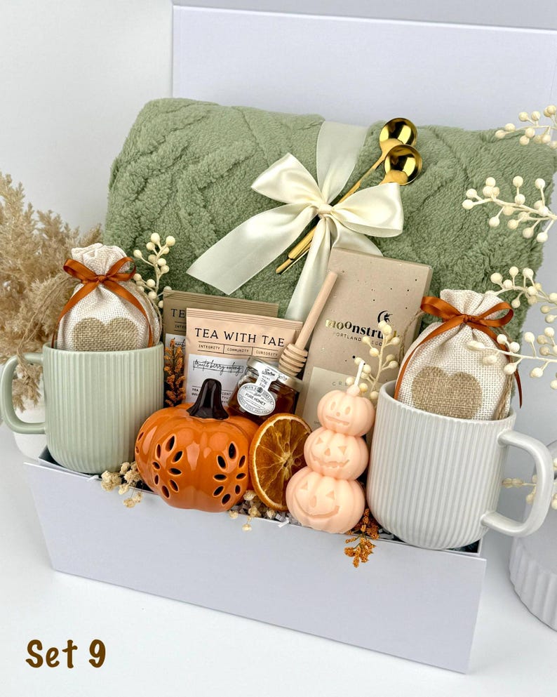 May include: A gift set in a white box containing a sage green textured throw blanket with a cream ribbon, two ribbed mugs, two heart-print pouches, a tea packet, a honey dipper, a small honey jar, a chocolate bar, a pumpkin candle, and dried fruit.
