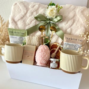 May include: A white gift basket filled with self-care items. Includes a plush cream-colored blanket, two cream-colored mugs, tea packets, soap, a decorative egg, honey, and a loofah. The basket is tied with a green ribbon and adorned with flowers.