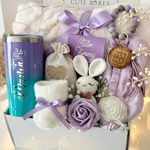 K&ouml;nnte beinhalten: Eine Geschenkbox mit verschiedenen Artikeln in Lila und Wei&szlig;. Enth&auml;lt einen Becher, eine Pl&uuml;schdecke, ein H&auml;schen aus H&auml;kelarbeit, einen Bei&szlig;ring und einen kleinen Beutel. Der Becher ist im Farbverlauf von T&uuml;rkis nach Lila mit dem Wort "Mama" versehen.