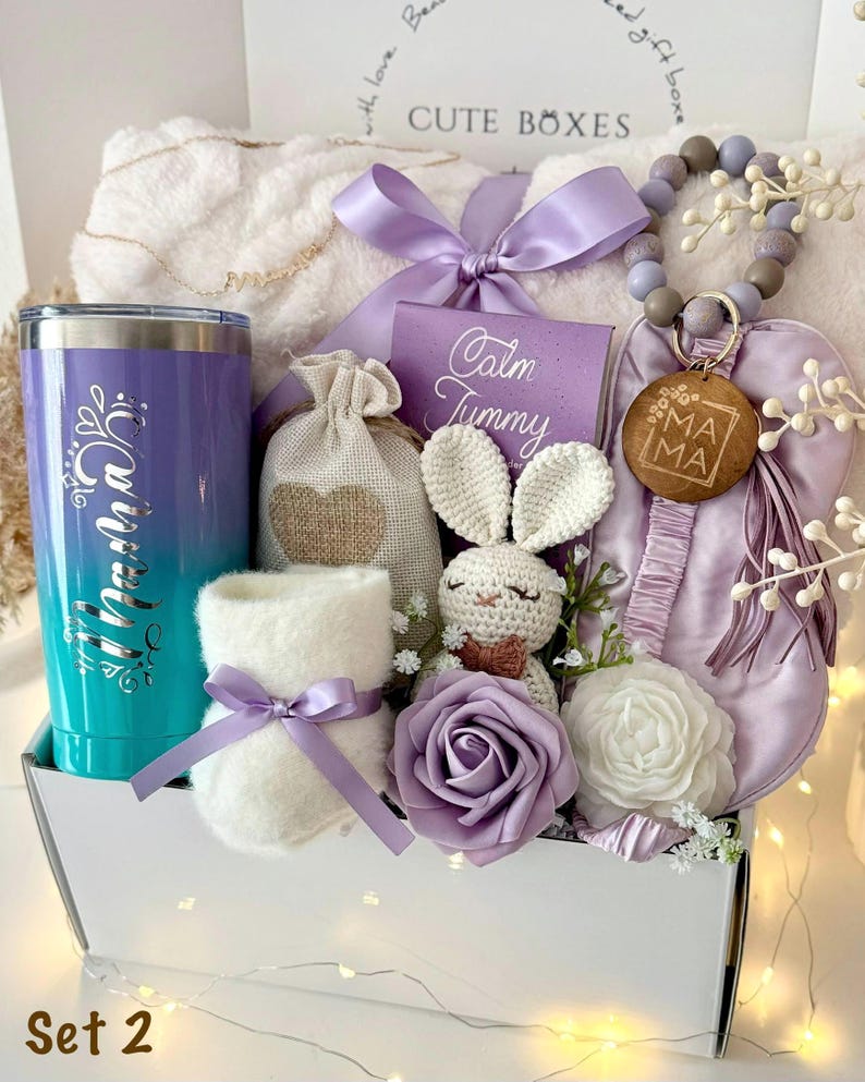 K&ouml;nnte beinhalten: Eine Geschenkbox mit verschiedenen Artikeln in Lila und Wei&szlig;. Enth&auml;lt einen Becher, eine Pl&uuml;schdecke, ein H&auml;schen aus H&auml;kelarbeit, einen Bei&szlig;ring und einen kleinen Beutel. Der Becher ist im Farbverlauf von T&uuml;rkis nach Lila mit dem Wort "Mama" versehen.
