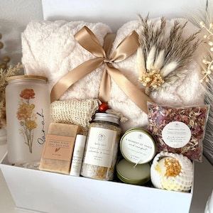 May include: A white gift box filled with self-care items. Includes a fluffy cream-colored blanket tied with a gold ribbon, a glass jar with floral design, soap, bath salts, a candle, and a bath bomb. The text "Set 8" is visible.