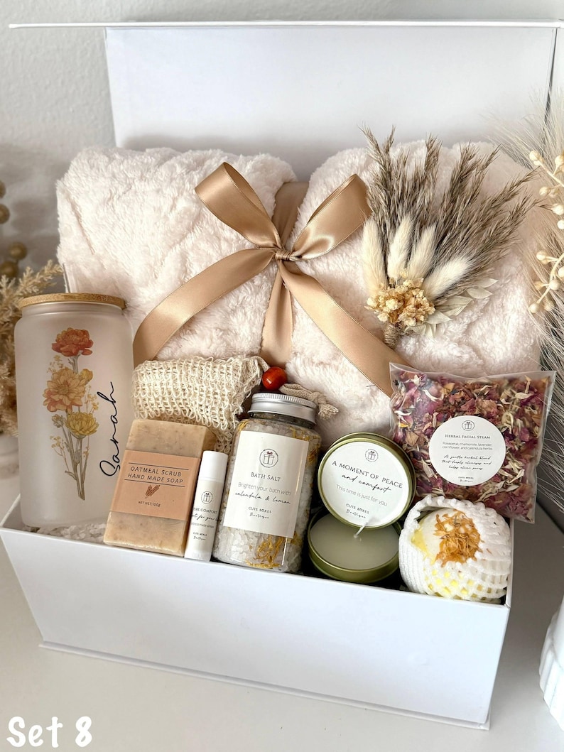May include: A white gift box filled with self-care items. Includes a fluffy cream-colored blanket tied with a gold ribbon, a glass jar with floral design, soap, bath salts, a candle, and a bath bomb. The text "Set 8" is visible.