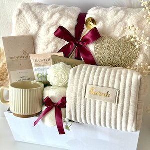 May include: A gift set containing a cream-colored cable knit blanket tied with a burgundy ribbon, a Moonstruck milk chocolate bar, a tea packet, a white rose-shaped candle, a ribbed mug, and a cream-colored corduroy pouch with the name Sarah.