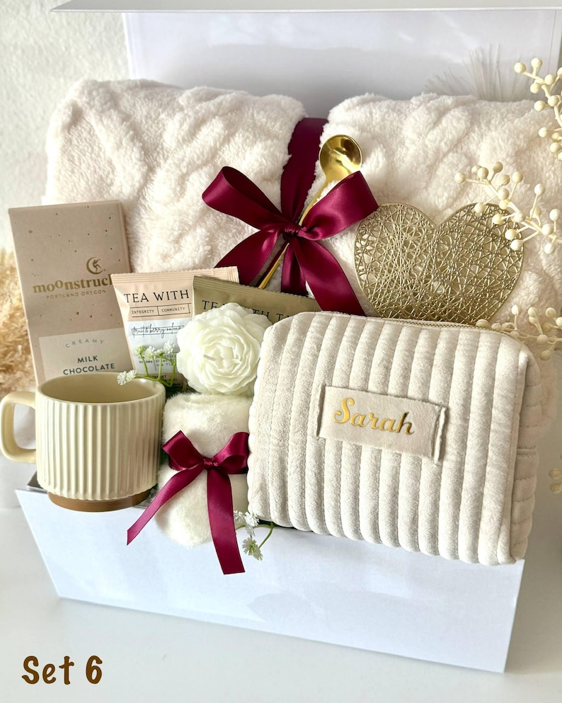 May include: A gift set containing a cream-colored cable knit blanket tied with a burgundy ribbon, a Moonstruck milk chocolate bar, a tea packet, a white rose-shaped candle, a ribbed mug, and a cream-colored corduroy pouch with the name Sarah.