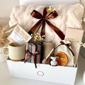 Puede incluir: Una cesta de regalo llena de una manta de felpa color crema, una taza, sobres de t&eacute;, velas, miel y una rodaja de naranja seca. La cesta est&aacute; atada con una cinta marr&oacute;n e incluye una cuchara dorada. El texto "Tea With Tae" es visible.