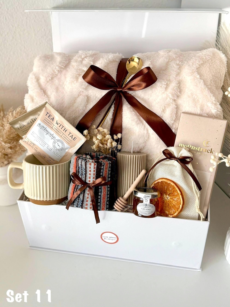 Puede incluir: Una cesta de regalo llena de una manta de felpa color crema, una taza, sobres de t&eacute;, velas, miel y una rodaja de naranja seca. La cesta est&aacute; atada con una cinta marr&oacute;n e incluye una cuchara dorada. El texto "Tea With Tae" es visible.