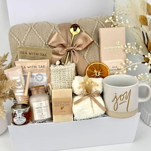 May include: A gift set in a white box containing a beige knit blanket with a satin ribbon, a gold spoon, a Moonstruck milk chocolate bar, a mug with the word joy, oatmeal soap, bath salts, honey, tea packets, a loofah, and a white fuzzy cloth.