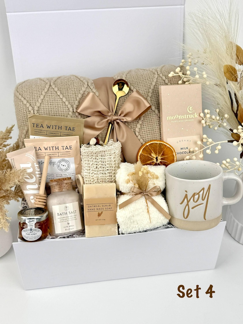 May include: A gift set in a white box containing a beige knit blanket with a satin ribbon, a gold spoon, a Moonstruck milk chocolate bar, a mug with the word joy, oatmeal soap, bath salts, honey, tea packets, a loofah, and a white fuzzy cloth.