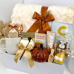 May include: A gift set in a white box containing a white ribbed mug, a plush white blanket with a brown ribbon, two tea packets, a Vitamin C mask, a shea butter tube, a small pumpkin-shaped decor, bath salts, honey, and a gold spoon.