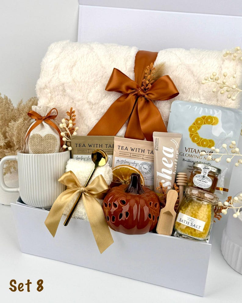 May include: A gift set in a white box containing a white ribbed mug, a plush white blanket with a brown ribbon, two tea packets, a Vitamin C mask, a shea butter tube, a small pumpkin-shaped decor, bath salts, honey, and a gold spoon.