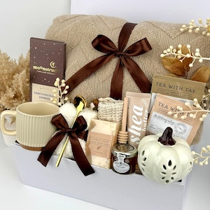 May include: A gift set in a white box containing a beige knit blanket with a brown ribbon, a Moonstruck dark chocolate bar, a ribbed beige mug, a gold spoon, oatmeal soap, a honey dipper, a jar of honey, shea butter, and a white ceramic pumpkin.