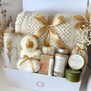 May include: A white gift box containing self-care products. It features a cream-coloured knitted blanket tied with a gold ribbon, a glass with floral design, bath salts, a candle, and other spa items.
