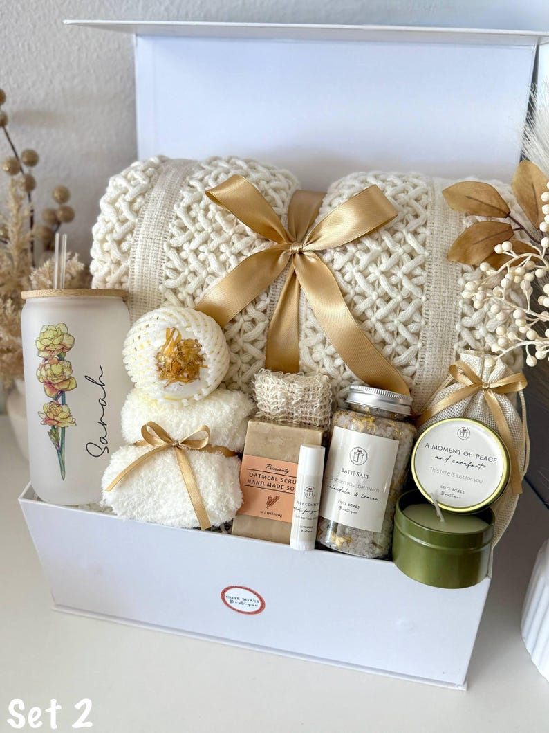 May include: A white gift box containing self-care products. It features a cream-coloured knitted blanket tied with a gold ribbon, a glass with floral design, bath salts, a candle, and other spa items.