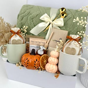 May include: A gift set in a white box containing a sage green textured throw blanket with a cream ribbon, two ribbed mugs, two heart-print pouches, a tea packet, a honey dipper, a small honey jar, a chocolate bar, a pumpkin candle, and dried fruit.