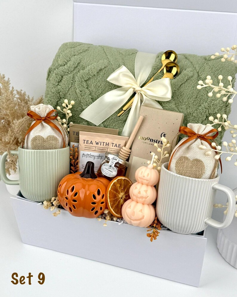 May include: A gift set in a white box containing a sage green textured throw blanket with a cream ribbon, two ribbed mugs, two heart-print pouches, a tea packet, a honey dipper, a small honey jar, a chocolate bar, a pumpkin candle, and dried fruit.