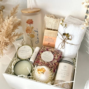 May include: A white gift box filled with self-care items. Includes a floral-decorated glass tumbler with a straw, a bar of oatmeal soap, a small white towel, bath salts, a candle, and a lip balm. The box is labeled "Set 9".