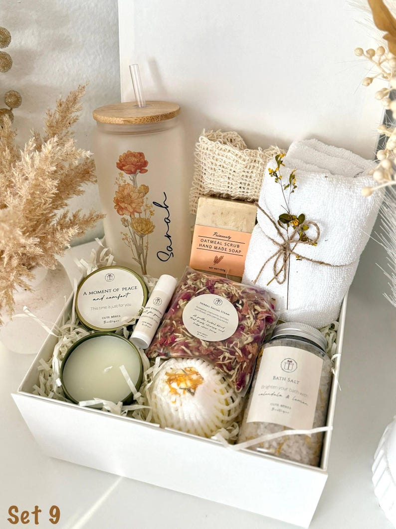 May include: A white gift box filled with self-care items. Includes a floral-decorated glass tumbler with a straw, a bar of oatmeal soap, a small white towel, bath salts, a candle, and a lip balm. The box is labeled "Set 9".