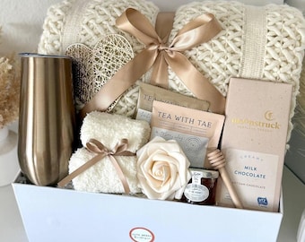 May include: A gift box containing a cream-coloured knitted blanket, a gold-coloured tumbler, a box of chocolate, tea packets, a small jar of honey, a decorative rose, and a small towel tied with a ribbon. The box is white with a gold ribbon.