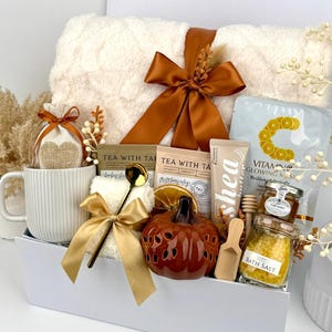 May include: A gift set in a white box containing a white ribbed mug, a plush white blanket with a brown ribbon, two tea packets, a Vitamin C mask, a shea butter tube, a small pumpkin-shaped decor, bath salts, honey, and a gold spoon.