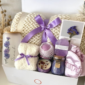May include: A white gift box containing self-care products. It features a cream-coloured pillow, a purple ribbon, a candle, a jar of bath salts, a small towel, a soap bar, and a satin sleep mask. The box also includes a glass with a floral design and the name "Sarah".