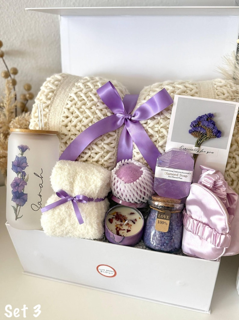 May include: A white gift box containing self-care products. It features a cream-coloured pillow, a purple ribbon, a candle, a jar of bath salts, a small towel, a soap bar, and a satin sleep mask. The box also includes a glass with a floral design and the name "Sarah".