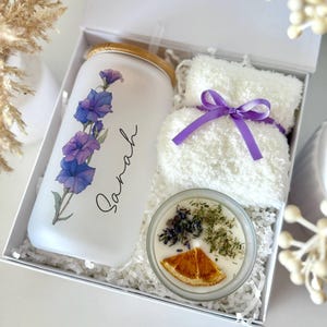 May include: A gift set in a white box includes a frosted glass tumbler with purple floral design and the name "Sarah", a white washcloth tied with a purple ribbon, and a candle with dried herbs and an orange slice.