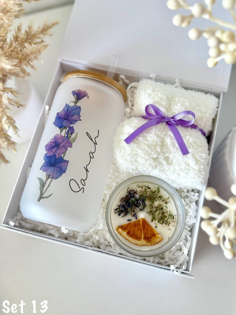 May include: A gift set in a white box includes a frosted glass tumbler with purple floral design and the name "Sarah", a white washcloth tied with a purple ribbon, and a candle with dried herbs and an orange slice.