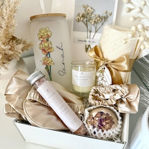 May include: A white gift box contains a tumbler with a floral design and the name "Sarah", a candle, bath salt, eye mask, scrunchies, and a bath bomb. The set is tied with a gold ribbon and includes a card with dried flowers.