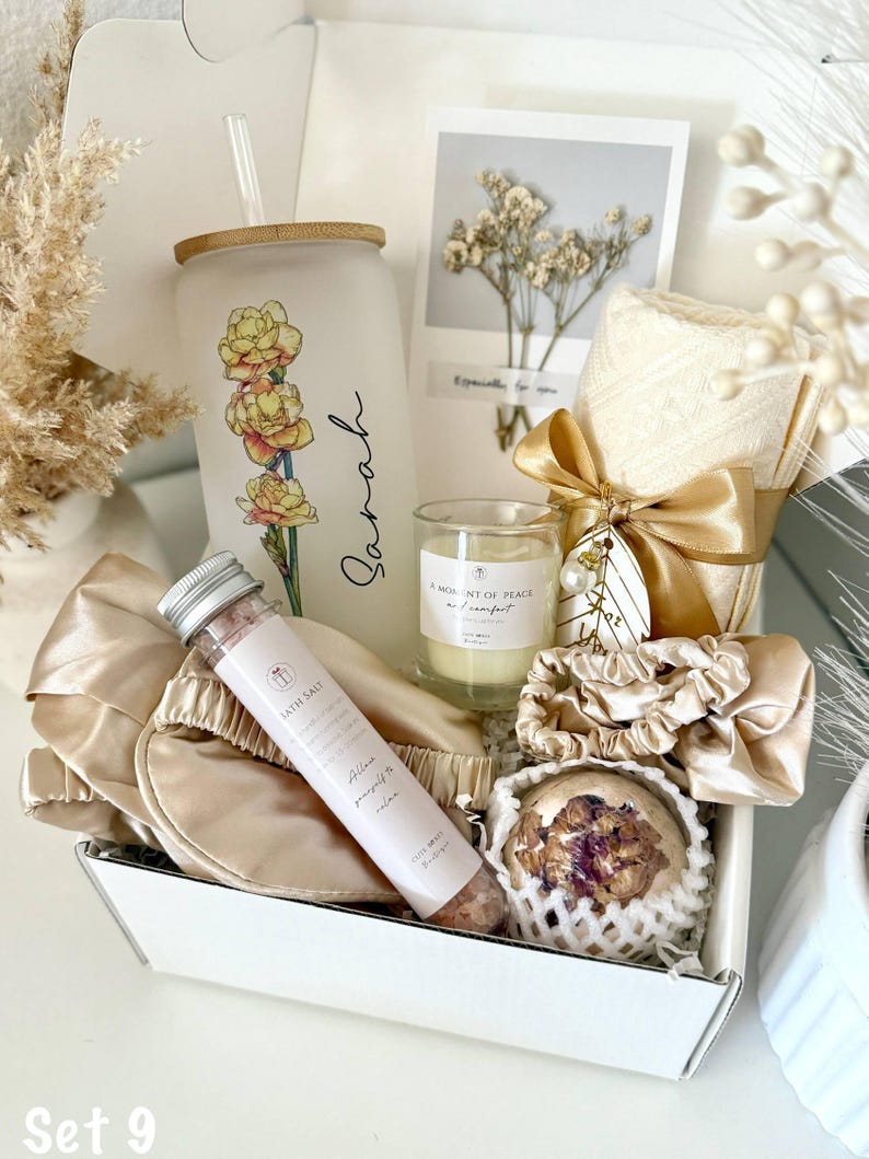 May include: A white gift box contains a tumbler with a floral design and the name "Sarah", a candle, bath salt, eye mask, scrunchies, and a bath bomb. The set is tied with a gold ribbon and includes a card with dried flowers.