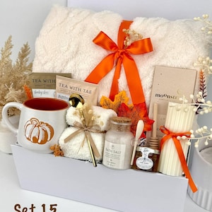May include: A gift set containing a white fuzzy blanket tied with an orange ribbon, a ceramic mug with a pumpkin graphic, two tea packets, a gold spoon, a small jar of bath salt, a jar of honey with a dipper, and a ribbed pillar candle.