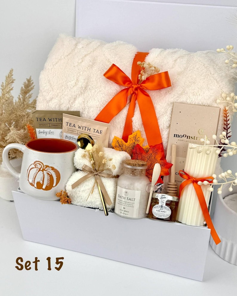 May include: A gift set containing a white fuzzy blanket tied with an orange ribbon, a ceramic mug with a pumpkin graphic, two tea packets, a gold spoon, a small jar of bath salt, a jar of honey with a dipper, and a ribbed pillar candle.