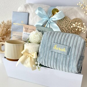 May include: A gift basket filled with a variety of items, including a light blue corduroy cosmetic bag with "Sarah" embroidered in gold, a cream-colored mug, tea, and a soft blanket. The basket is tied with a light blue ribbon and includes decorative flowers.