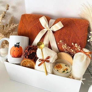 May include: A white gift box filled with a burnt orange towel tied with a cream ribbon, a mug with a pumpkin illustration, a small candle, and other spa-themed items. A gold spoon and dried orange slices are also included.