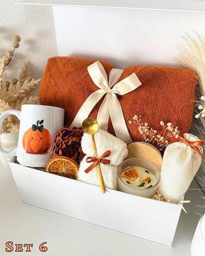 May include: A white gift box filled with a burnt orange towel tied with a cream ribbon, a mug with a pumpkin illustration, a small candle, and other spa-themed items. A gold spoon and dried orange slices are also included.