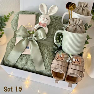 May include: A gift set containing a sage green plush blanket tied with a satin ribbon, a crochet bunny rattle, a light green ribbed ceramic mug with a burlap pouch, two gold spoons, and a pair of tan corduroy baby shoes with bear faces.