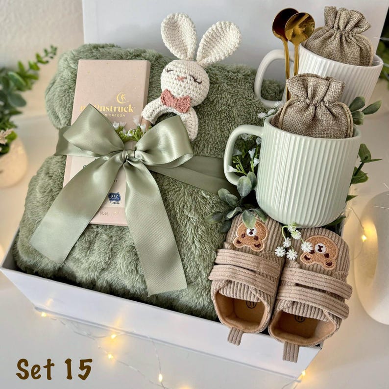 May include: A gift set containing a sage green plush blanket tied with a satin ribbon, a crochet bunny rattle, a light green ribbed ceramic mug with a burlap pouch, two gold spoons, and a pair of tan corduroy baby shoes with bear faces.