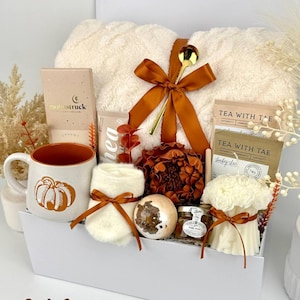 May include: A gift box containing a cream-colored fuzzy blanket tied with a brown ribbon and gold spoon, a mug with a pumpkin graphic, a small jar of honey, a bath bomb, a white candle, tea packets, a chocolate bar, and a pair of cream socks.