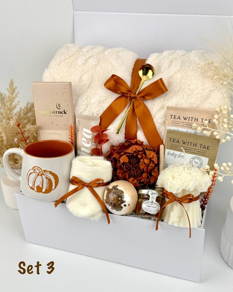 May include: A gift box containing a cream-colored fuzzy blanket tied with a brown ribbon and gold spoon, a mug with a pumpkin graphic, a small jar of honey, a bath bomb, a white candle, tea packets, a chocolate bar, and a pair of cream socks.