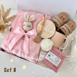 May include: A gift set containing a soft pink plush blanket tied with a satin ribbon, a crocheted bunny rattle, a wooden pacifier clip with peach beads, a round wooden sign reading hello world, beige baby booties with bear faces, and tea.