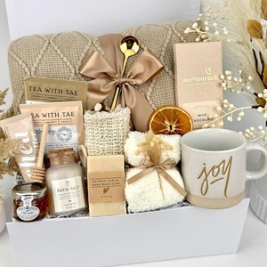 May include: A gift box containing a beige knit blanket with a gold ribbon, a gold spoon, Moonstruck milk chocolate, Tea with Tae tea bags, a honey jar with a dipper, bath salt, oatmeal soap, a loofah, a white mug with the word joy, and dried fruit.