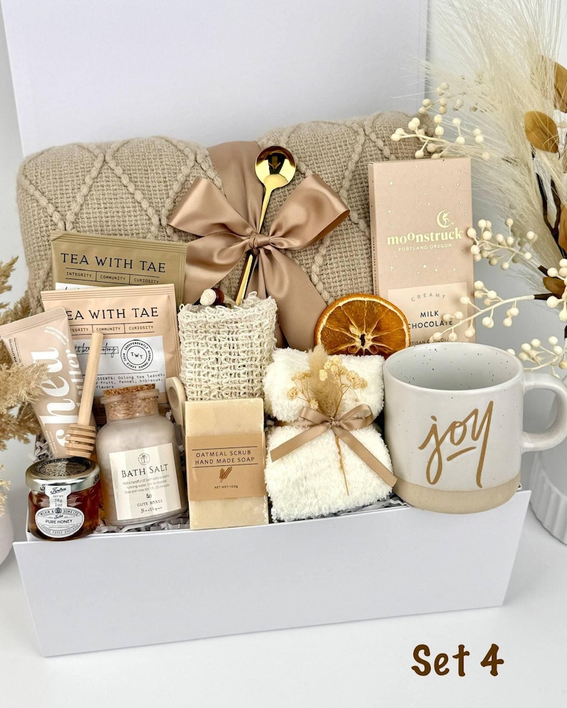 May include: A gift box containing a beige knit blanket with a gold ribbon, a gold spoon, Moonstruck milk chocolate, Tea with Tae tea bags, a honey jar with a dipper, bath salt, oatmeal soap, a loofah, a white mug with the word joy, and dried fruit.