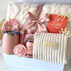 May include: A gift box filled with self-care items. Includes a fluffy white blanket tied with a pink ribbon, a pink tumbler, a candle, a sleep mask, a scrunchie, a rose, and a tea packet. A beige pouch with the name "Sarah" is also included.
